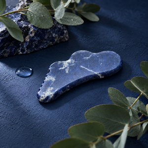 A Cecily Braden Sodalite stone gua sha massage tool with natural white veining, displayed on a textured dark blue surface with eucalyptus leaves and a small water droplet.