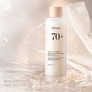 Anua 70+ Rice 70 + Ceramide Milky Hydration bottle with a pale peach cap, styled on a reflective glass surface with soft translucent paper and pearl props.