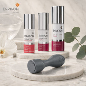 Three ENVIRON Focus Care + serum bottles in white and silver packaging with pink-red bases, displayed on a marble pedestal alongside a grey Environ Cosmetic Focus CIT with a glass bowl, white flower, and eucalyptus leaves in a bright, clean setting.