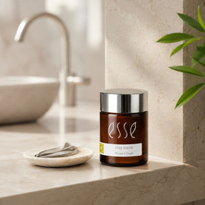 An amber glass jar of Esse Clay Mask with a silver lid sits on a beige stone countertop next to a small dish with a folded towel. The background shows a softly blurred bathroom sink and faucet, with a green plant leaf visible on the right.