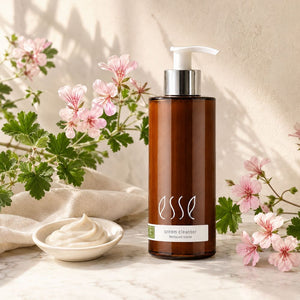 An amber glass pump bottle of ESSE Cream Cleanser moisturising cleanser sits on a marble surface, surrounded by pink flowers, green leaves, a beige cloth, and a small dish of cream.