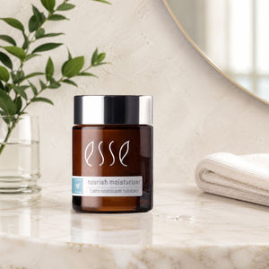A jar of Esse Nourish Moisturiser sits on a polished marble countertop in a bathroom setting, flanked by a glass vase with green leaves on the left and a folded white towel on the right.