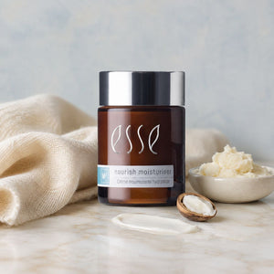 Essé nourish moisturiser cream in an amber glass jar with a silver lid, styled on a marble surface with a beige cloth, a bowl of cream, and a Shea Butter Nut.