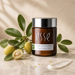 A jar of Esse Rich Moisturiser skincare cream placed on a marble surface with olive branches and a smear of cream beside it, against a softly lit beige background.