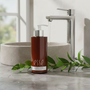 A bottle of Esse Sensitive Cleanser stands on a marble countertop in front of a running faucet and stone sink, with green leaves placed around the bottle.