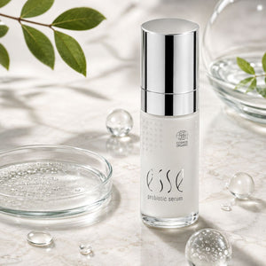 Organic ESSE Skincare UK Probiotic Serum in a clear bottle with a reflective silver cap, set on a marble surface with glass dishes, clear liquid droplets, and green leaves.