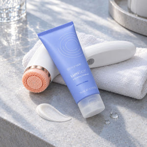 A tube of Nu Skin AgeLoc LumiSpa activating cleanser in a light blue squeeze packaging, displayed on a gray stone countertop next to a white and rose gold facial cleansing device, a white towel, and a smear of cleansed cream.