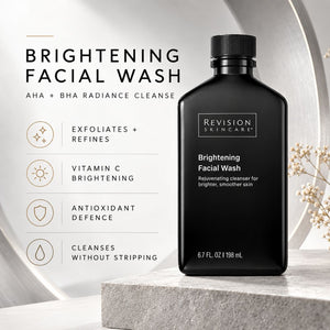 Revision Skincare Brightening Facial Wash in a matte black bottle, displayed on a stone pedestal next to product benefits text on a light gray background.
