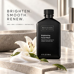 Revision Skincare Brightening Facial Wash in a matte black bottle, styled on a stone surface with a white lily flower and green leaves, alongside the brand's brightening slogan.