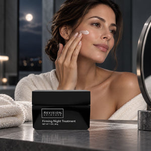 A woman in a white robe applies cream to her cheek while looking into a mirror, with a black Revision Skincare jar labeled 'Firming Night Treatment' on a bathroom counter in the foreground.