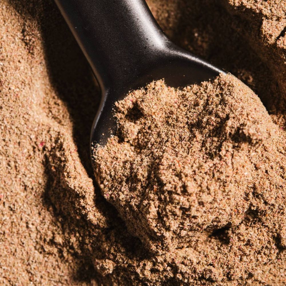 A close-up shot of a black scoop filled with Skin Integrate 28 Biome powder, set against a background of the same brown powder.