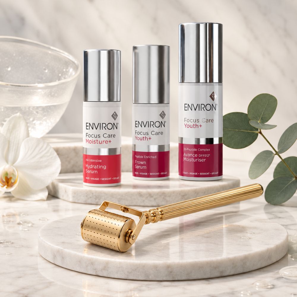 Three ENVIRON Focus Care youth+ serum bottles in white and silver packaging with magenta-red labels, displayed on a marble pedestal alongside a gold facial roller tool, with decorative eucalyptus leaves and a glass bowl in the background.