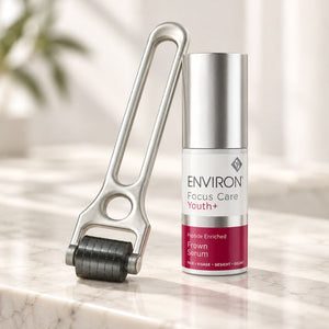 A silver derma roller tool and a bottle of ENVIRON Focus Care Youth+ Frown Serum placed on a light marble surface with a softly blurred indoor background.