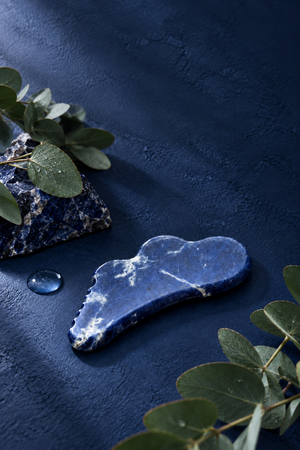 A Cecily Braden Sodalite Blue stone gua sha massage tool with white veining, displayed on a dark textured blue surface with eucalyptus leaves and a small water droplet.