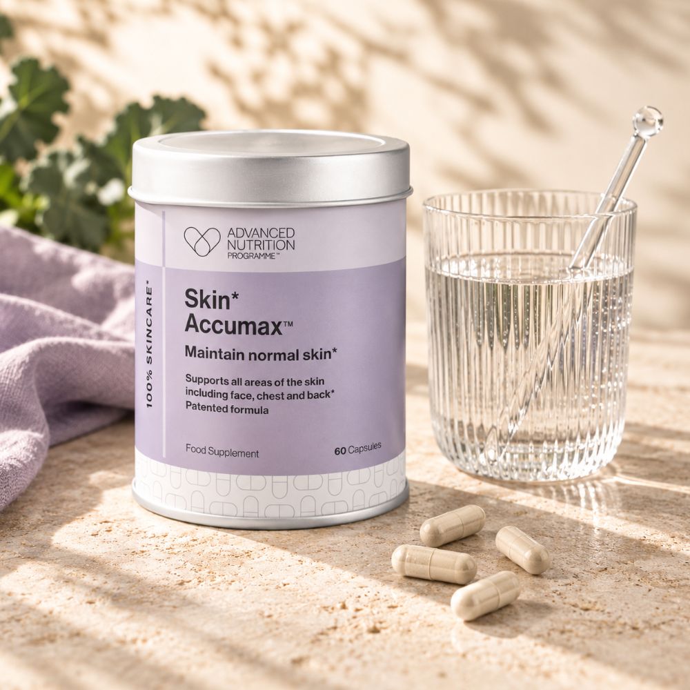 A white supplement jar labeled 'Skin+ Accumax' stands on a sunlit stone surface beside a glass of water with a spoon and a few capsules scattered in front. The scene includes soft shadows and a purple cloth, making it a styled product setup rather than a plain studio shot.