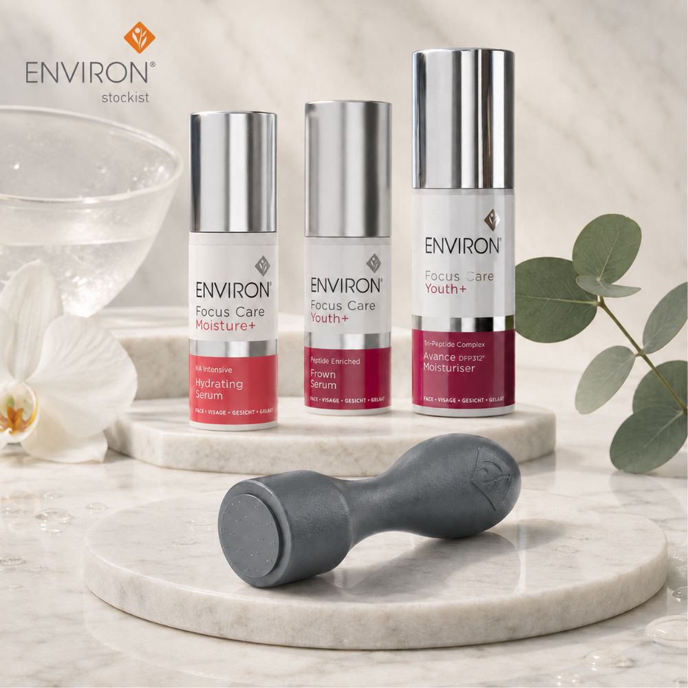 Three ENVIRON Focus Care + serum bottles in white and silver packaging with pink-red bases, displayed on a marble pedestal alongside a grey Environ Cosmetic Focus CIT with a glass bowl, white flower, and eucalyptus leaves in a bright, clean setting.