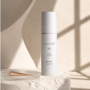 A white cylindrical bottle of EPICUTIS Lipid Shield Sunscreen SPF 50 sunscreen, displayed on a textured beige stone pedestal with a smear of beige foundation in a clear dish. The background is a warm beige wall with soft window shadow patterns.