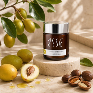 A jar of ESSE Cocoa Exfoliator sits on a stone pedestal, surrounded by green mangoes, mango halves, and cashews on a warm beige stone surface with a leafy branch in the background.