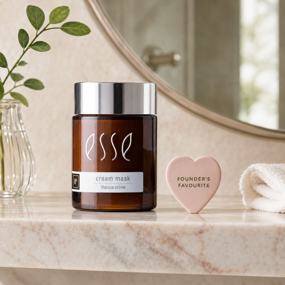 Esse cream mask jar in an amber glass bottle with a silver lid, set on a marble countertop next to a pink heart-shaped soap and a rolled white towel, with a mirror and plant in the background.