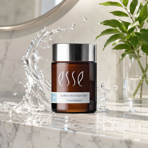 Essé Hydro Moisturiser cream jar in an amber brown bottle with a silver cap, set on a marble countertop with a water splash and a green plant in the background.