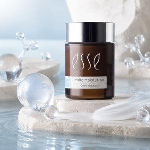 Essé Hydro Moisturiser cream jar in a dark brown bottle with a silver cap, styled on a light stone platform with clear glass spheres and water.
