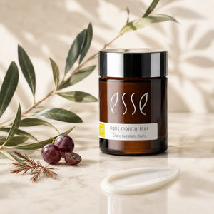 Essé Light Moisturiser cream jar in an amber glass bottle with a black lid, styled on a marble surface with olive branches, olives, and a white silicone applicator.