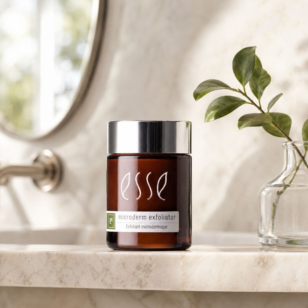 An amber glass jar of Esse Microderm Exfoliator with a shiny silver cap, set on a marble countertop next to a glass vase with green leaves.