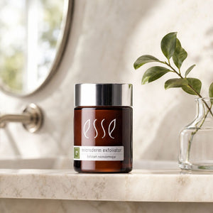 An amber glass jar of Esse Microderm Exfoliator with a shiny silver cap, set on a marble countertop next to a glass vase with green leaves.