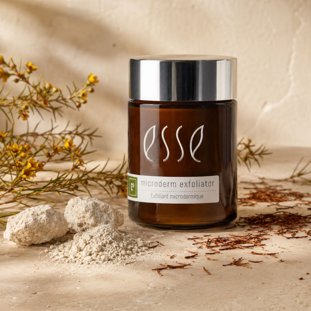 A jar of ESSE microderm exfoliator with a silver lid and amber glass bottle, styled on a beige surface with dried flowers, loose exfoliating powder, and small stones.