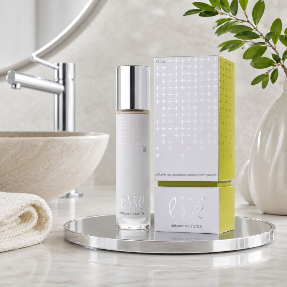 ESSE Defence Moisturiser bottle and packaging box displayed on a round silver tray in a bathroom setting with a sink, mirror, and plant visible in the background.