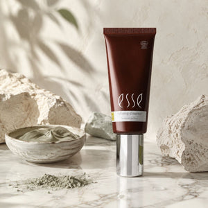 A squeeze tube of Esse Refining Cleanser stands upright on a marble surface, surrounded by textured stones and a small bowl of green cleanser cream. The tube is dark brown with a reflective silver cap.