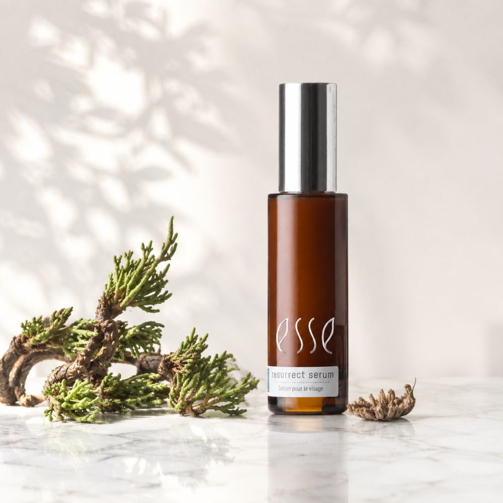 Essence Resurfact Serum in an amber glass bottle with a silver cap, styled on a marble surface with green botanical elements against a softly lit background.
