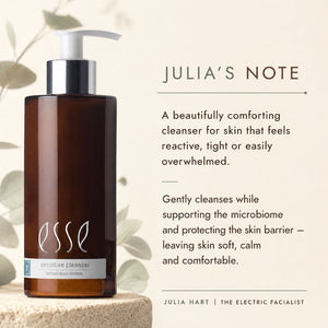 Esse Sensitive Cleanser bottle in a dark amber brown glass with a white pump and silver collar, displayed on a stone pedestal next to descriptive text about 'Julia's Note' on a warm beige background with soft leaf illustrations.