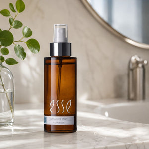 An amber glass spray bottle of Esse Sensitive Mist with a black pump and clear cap, standing on a white marble countertop in a bathroom setting with a plant and mirror in the background.