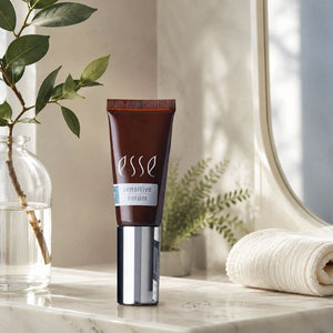 A squeeze tube of Esse Sensitive Serum stands on a marble surface in a softly styled bathroom setting. The tube is dark brown with a reflective silver cap, and the background includes a glass vase with green leaves, a rolled cream towel, and warm natural light.