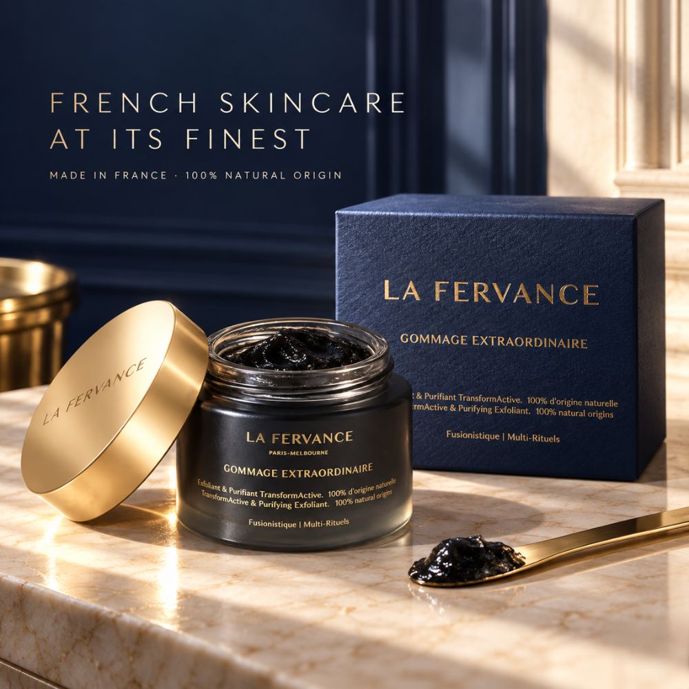 La Fervance Gommage Extraordinaire facial scrub in an open dark glass jar with a gold lid, displayed on a marble countertop next to its navy blue box and a gold spoon holding some of the black scrub.