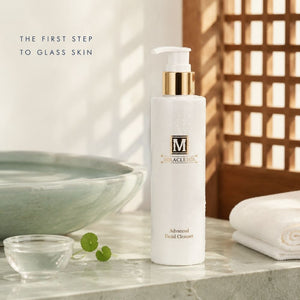 A bottle of Miracletox Advanced Facial Cleanser stands on a marble surface in a Korean bathroom-like setting with a bowl, small glass dish, Centella leaves, and a folded towel. The bottle is white with a gold pump collar and a white pump top.