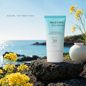 A tube of K-BeautyMiracletox Mild Cool Sun Cream stands on a dark rock with a scenic ocean and coastal landscape in the background, framed by yellow flowers. The product packaging is light blue and white, with text indicating SPF 50+ protection.