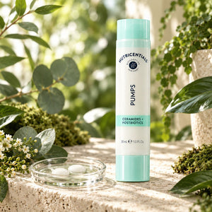 A 30ml bottle of Nu Skin Nutricentials  Ceramides + Postbiotics PUMPS, styled on a stone surface with green foliage and a small glass dish in a garden setting.