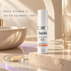 A bottle of Senté Defense C Serum stands on a beige stone slab beside an open clear lid containing yellow serum, with a ceramic bowl and glass vessel in the background. Text at the top reads 'DAILY VITAMIN C' and '20% THD ASCORBATE + HSA'.