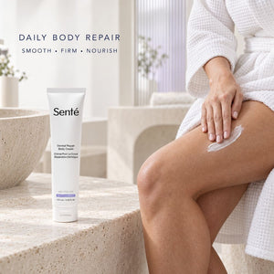 A tube of Sente Dermal Repair Body Cream stands on a stone surface beside a woman in a white robe sitting on a bench, with cream smeared on her thigh. The scene is softly lit and styled in a bathroom-like setting.